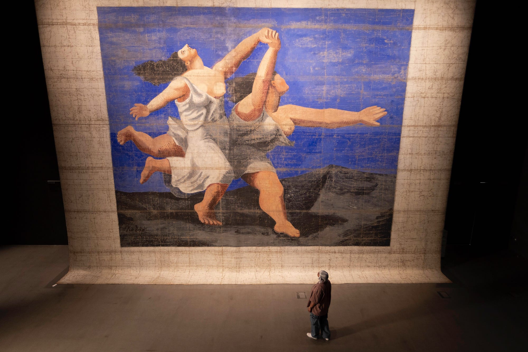 a figure stands in front of a giant Picasso mural of two frolicking figures on blue background